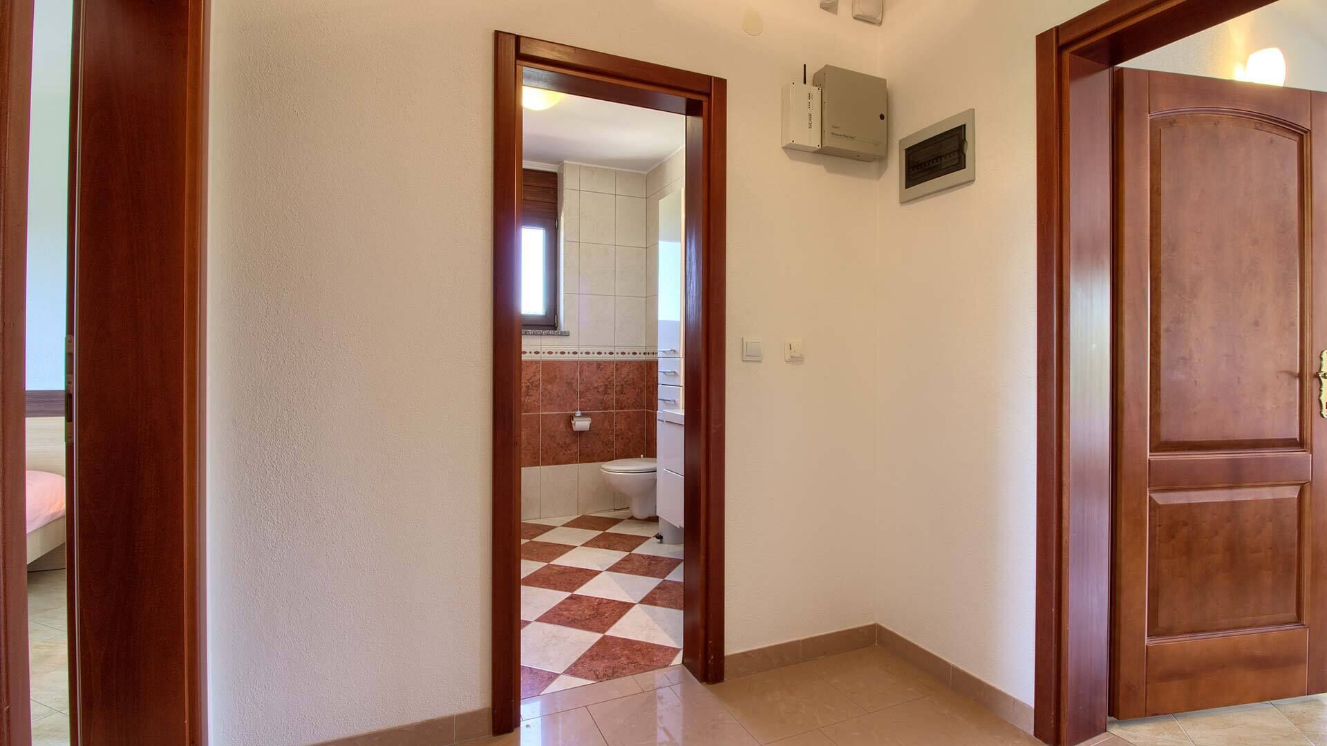 Ein Flur mit Schachbrettfußboden, einer Holztür und Blick ins Badezimmer mit Toilette und gefliesten Wänden.