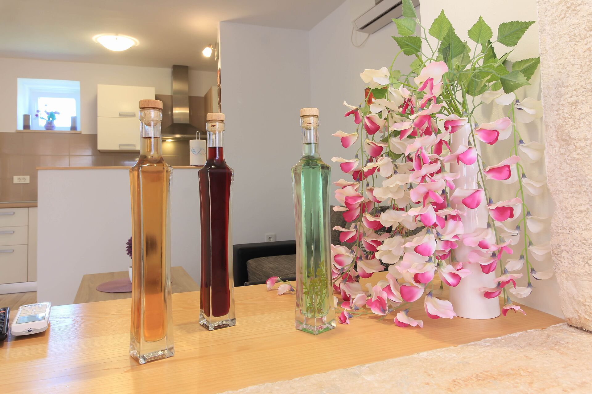 Three bottles of liquid on a wooden table, a white vase with pink and white flowers, and a kitchen area in the background.