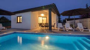 A swimming pool with lounge chairs and umbrellas, a patio area with a table and chairs, and a building with a tiled roof.
