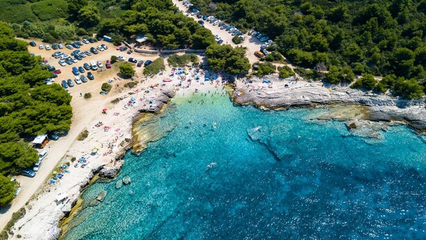 An aerial view of a coastal area with a beach, clear blue waters, and a parking lot with cars.