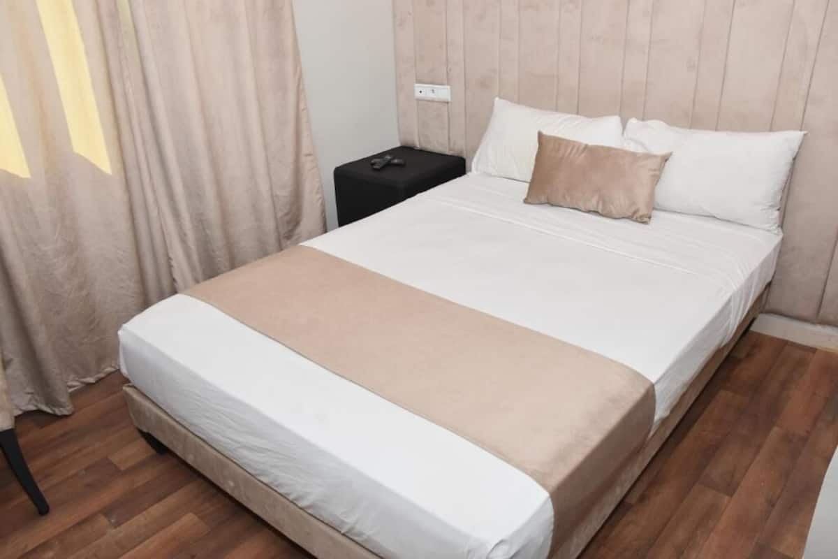A neatly made bed with a white and beige bedspread, a black nightstand with a lamp, and a wooden headboard.