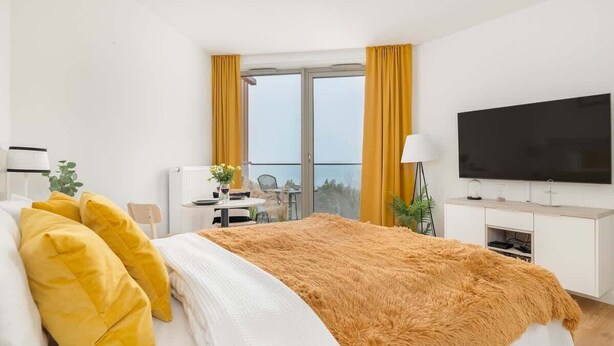 A modern hotel room with a large bed, a flat-screen TV, a desk with a chair, and a balcony with yellow curtains. - Studio with Sea View | Parking | By the Beach (Pobierowo)