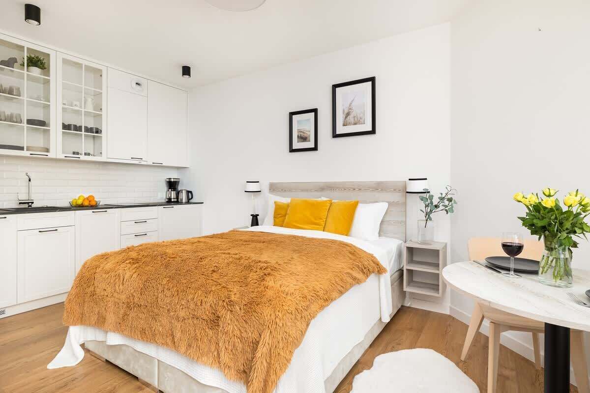 A modern hotel room with a bed, a small dining table, a kitchen area, and a white color scheme.