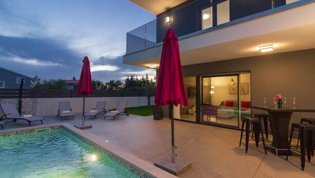 A modern outdoor pool area with lounge chairs and umbrellas, a dining table with chairs, and a glass-enclosed patio. - Villa Ema with private pool by WiiBuk (Istria - Sisan) (Sisan)