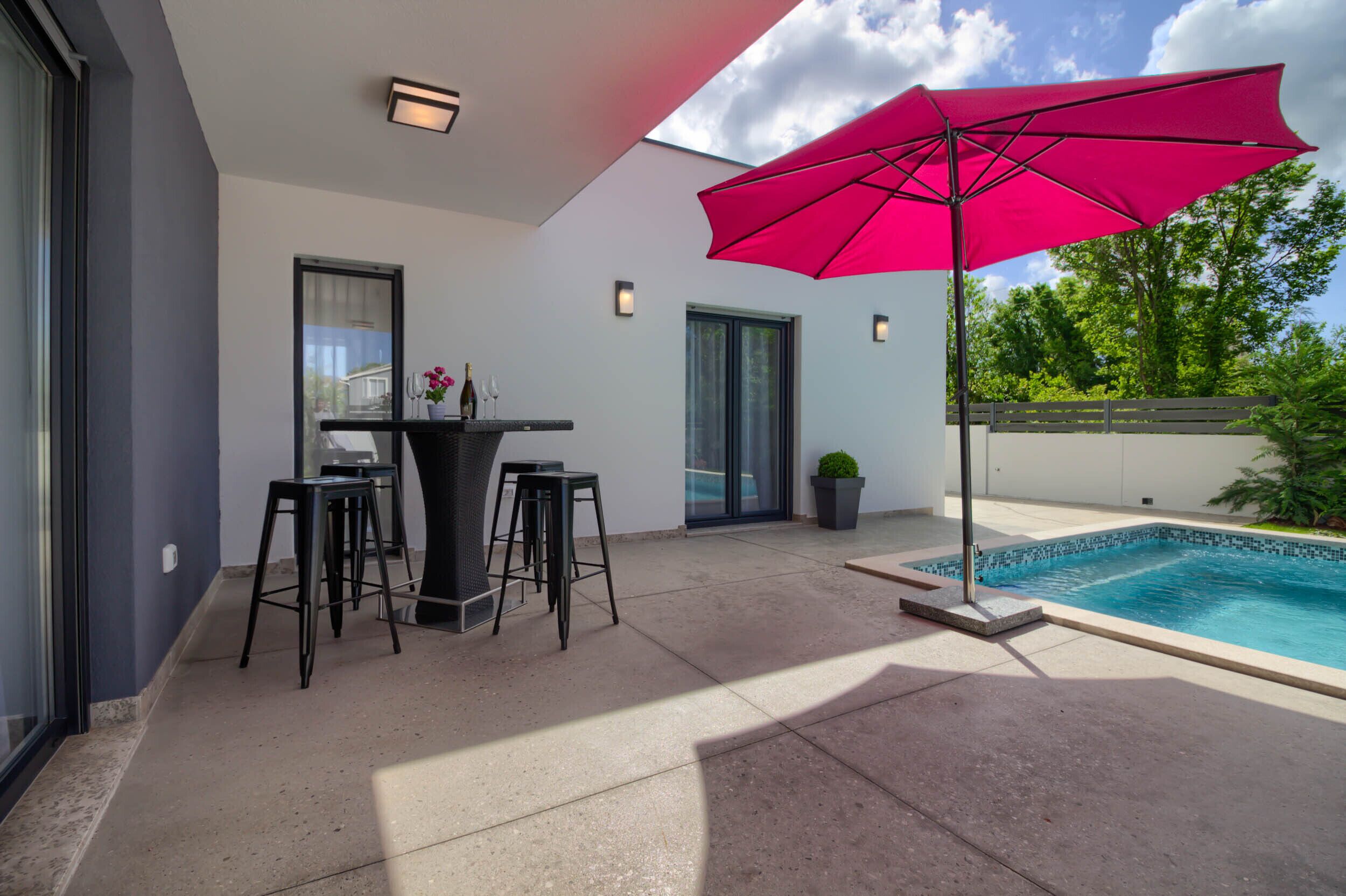 A modern outdoor patio with a pool, a table with stools, and a large pink umbrella.