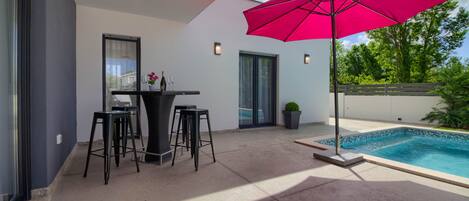 A modern outdoor patio with a pool, a table with stools, and a large pink umbrella.