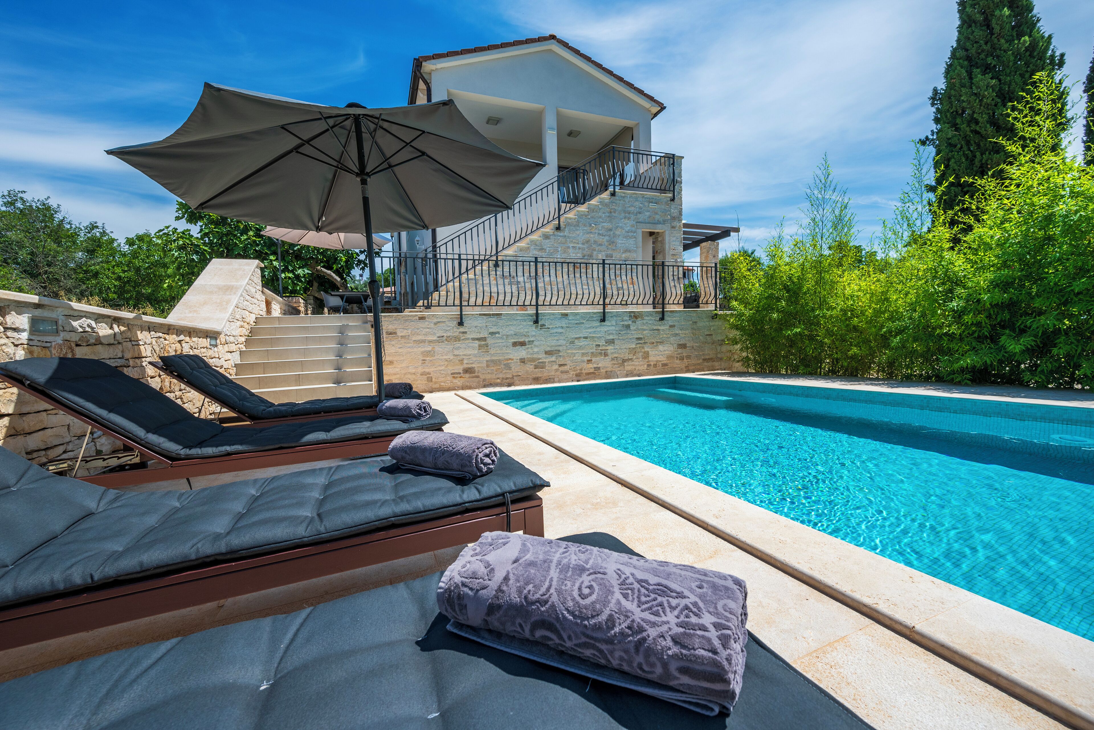 A poolside area with lounge chairs, a large umbrella, and a stone staircase leading to a covered balcony.