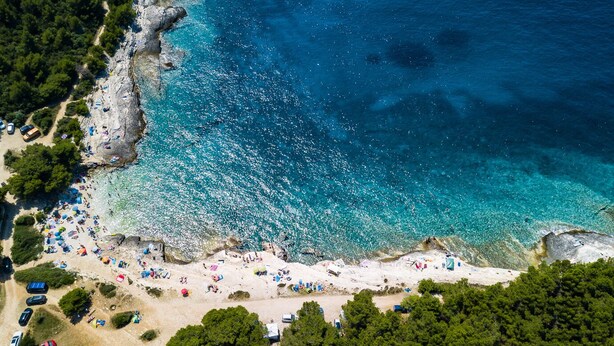 Vista aerea di una spiaggia con acque turchesi cristalline, costa rocciosa e bagnanti sparsi.