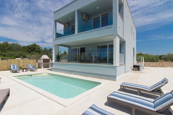 Une maison moderne à deux étages, dotée d’une piscine, d’un espace de détente extérieur et offrant une vue sur la mer.