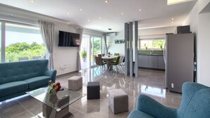 A modern living room with a sofa, ottomans, a glass coffee table, and a flat-screen TV. There is a dining area with a table and chairs, and a kitchen with a refrigerator. - Villa Horizont (Istria - Fažana) (Fažana)