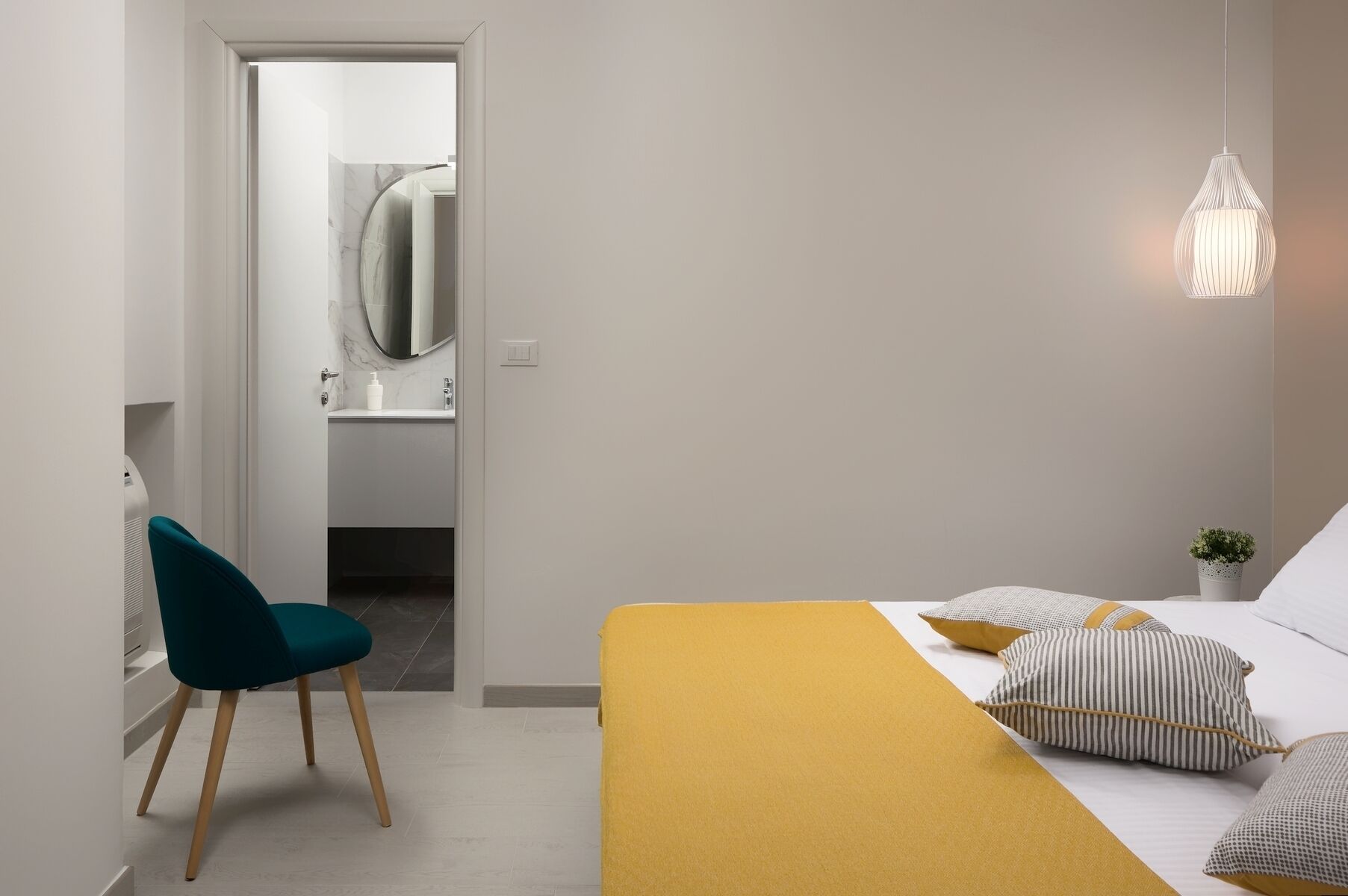 A bedroom with a bed, a chair, a bedside table with a plant, and a hanging light fixture.