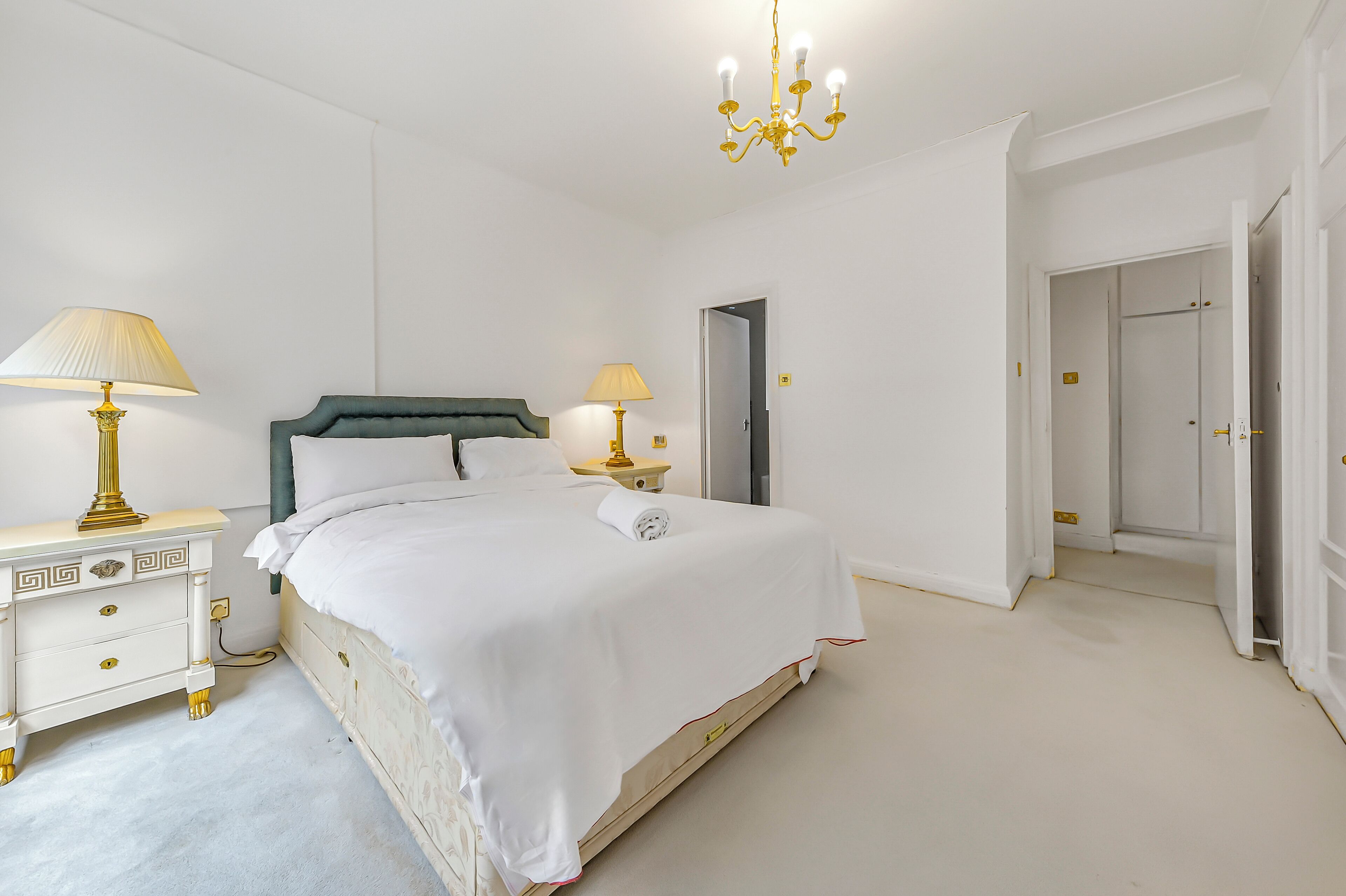 A neatly arranged bedroom with a large bed, two bedside tables with lamps, a chandelier, and a door leading to another room.