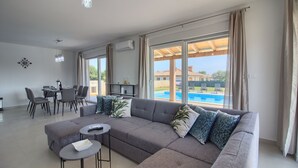 Ein modernes Wohnzimmer mit einem grauen Ecksofa, zwei kleinen runden Tischen, einem Essbereich mit Stühlen und einem großen Fenster mit Blick auf einen Swimmingpool.