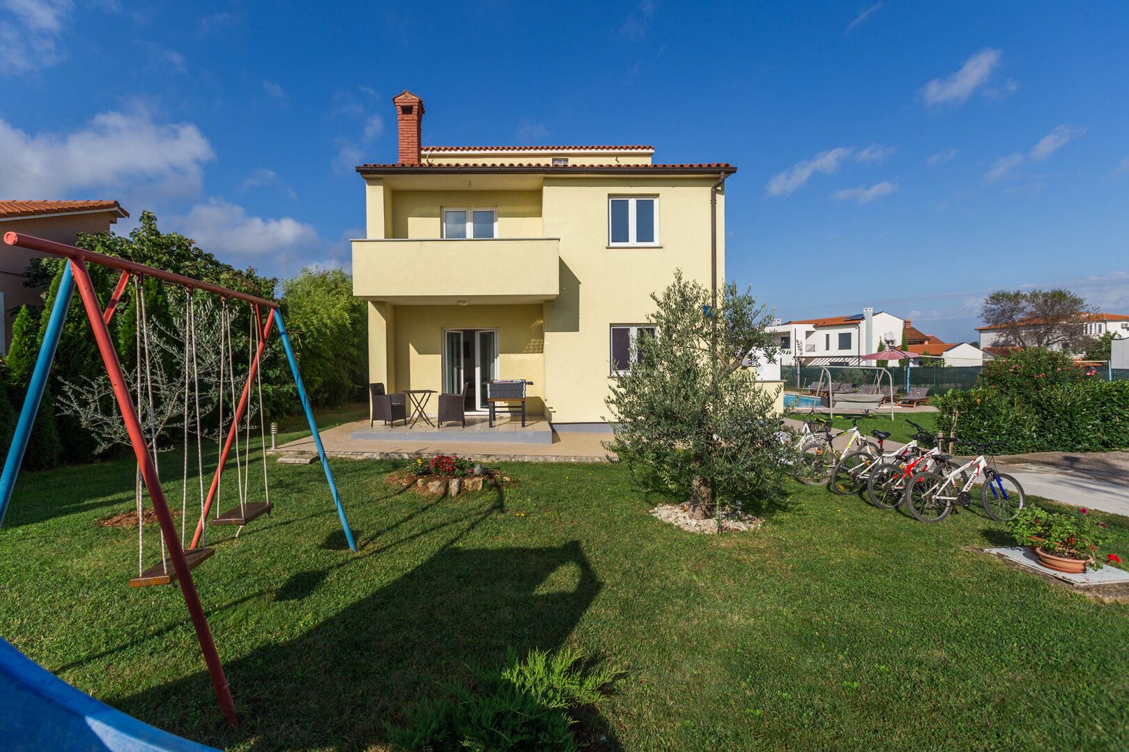 Une maison à deux étages avec un patio, une balançoire et un petit arbre dans le jardin.