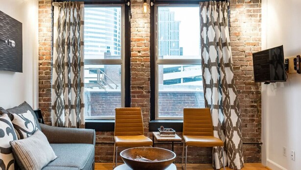 A modern living room with a brick wall, two large windows with patterned curtains, a grey sofa, two brown chairs, a wooden coffee table, and a flat-screen TV mounted on the wall.
