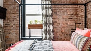 A bedroom with a brick wall, a window with a view, a bed with patterned bedding, and a plant on the windowsill.