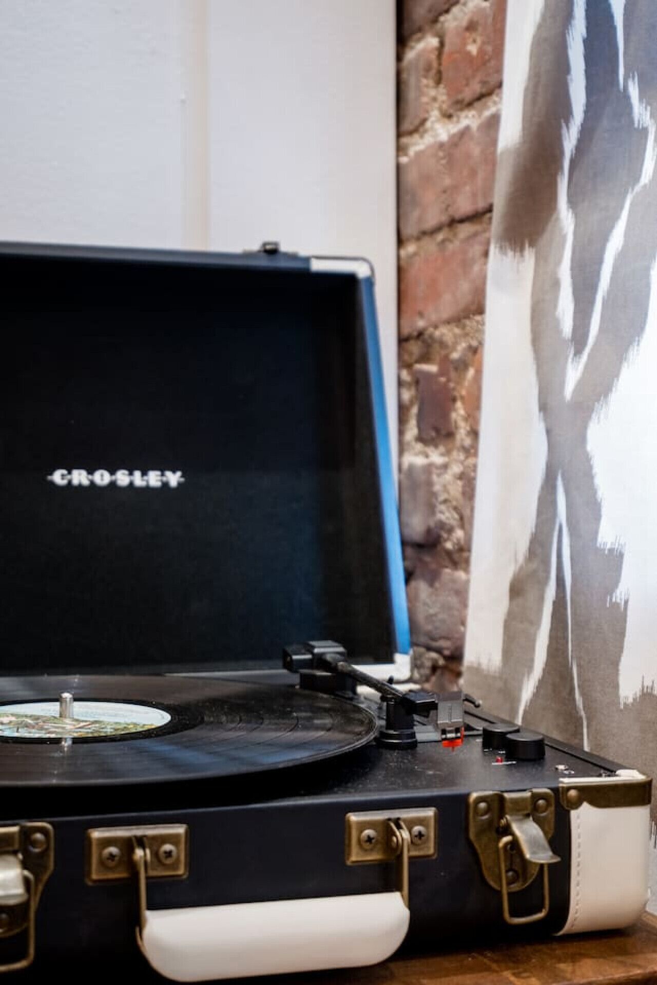 A Crosley turntable in a black and white case with brass accents, placed on a wooden surface against a brick wall and a patterned curtain.
