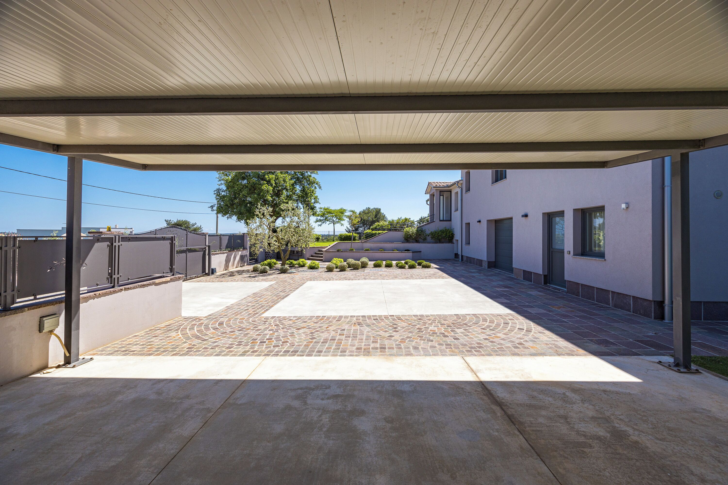 A covered outdoor area with a paved walkway, a small garden with trimmed bushes, and a building with a modern design in the background.