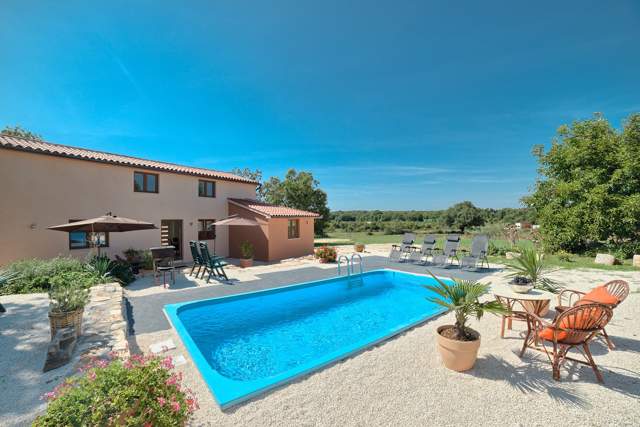 A swimming pool with lounge chairs and a table, surrounded by potted plants and a building with a tiled roof.
