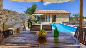 Une table en bois avec un saladier de fruits et une bouteille verte, un mur en pierre, un évier en pierre et un espace piscine avec des chaises longues.