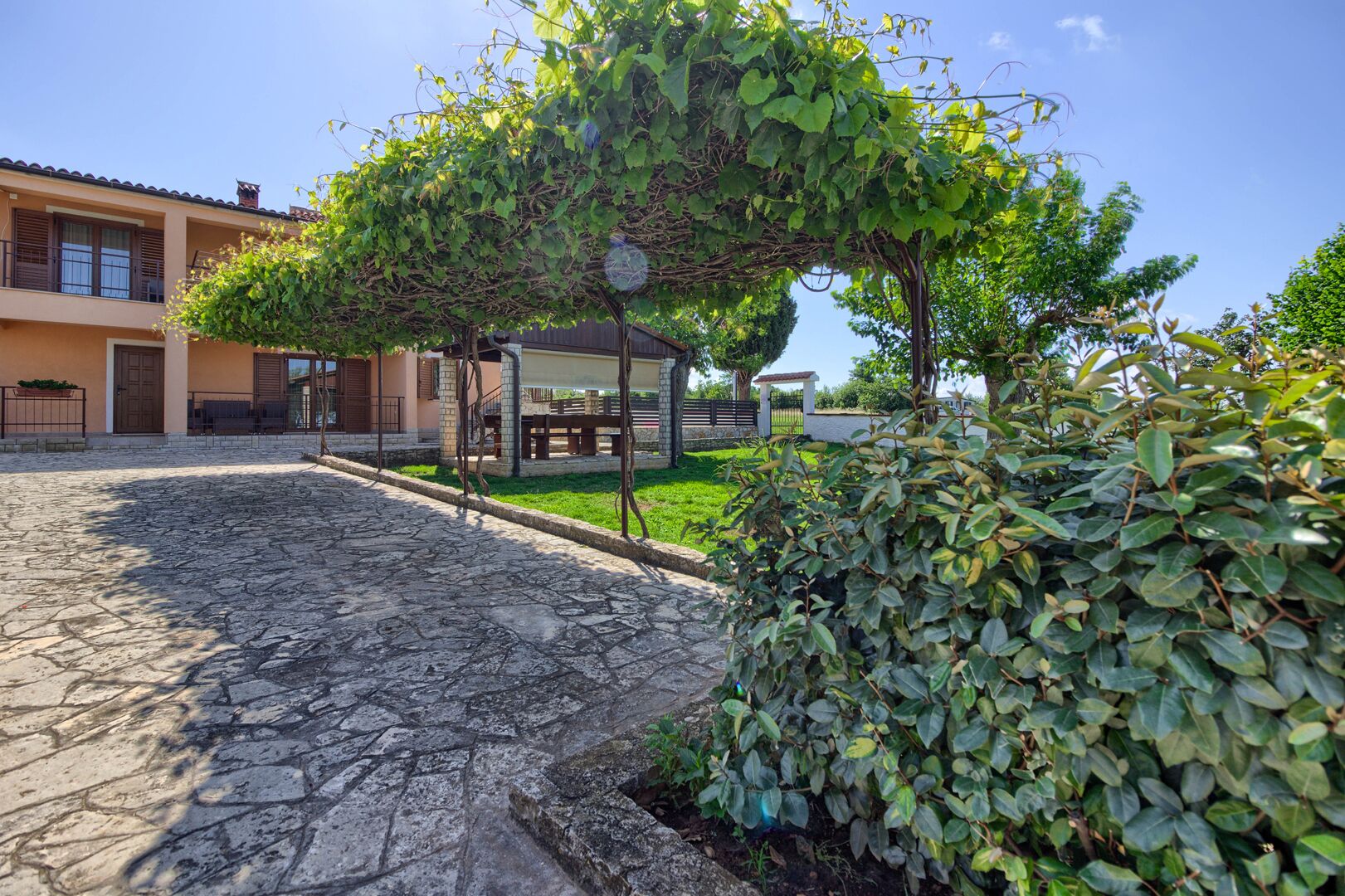 A courtyard with a stone pathway, a pergola covered with climbing plants, a small seating area, and a building with a tiled roof.