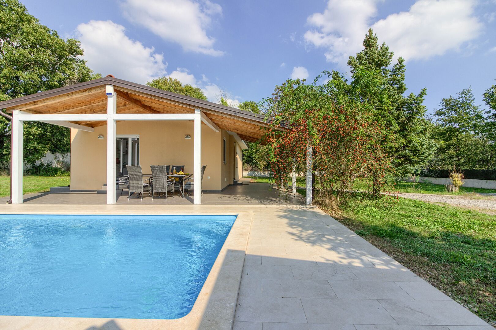 A covered outdoor area with a pool, a patio with chairs, and a small building with a sloped roof.