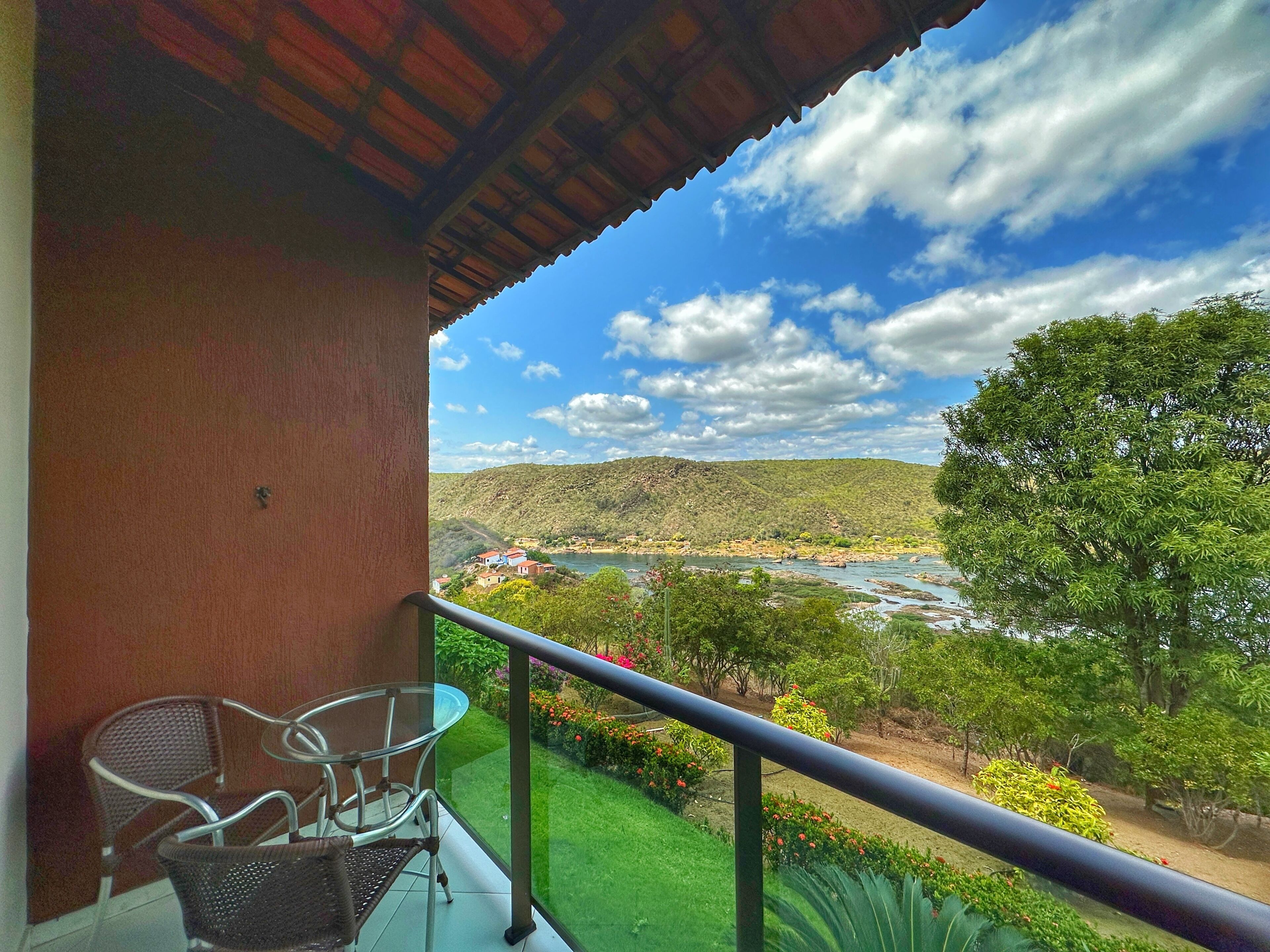 A balcony with a glass table and chairs, overlooking a river and hilly landscape.