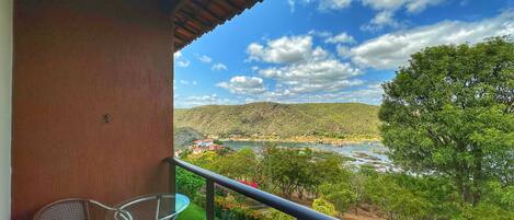 Un balcón con mesa y sillas de vidrio, con vista a un río y un paisaje montañoso.