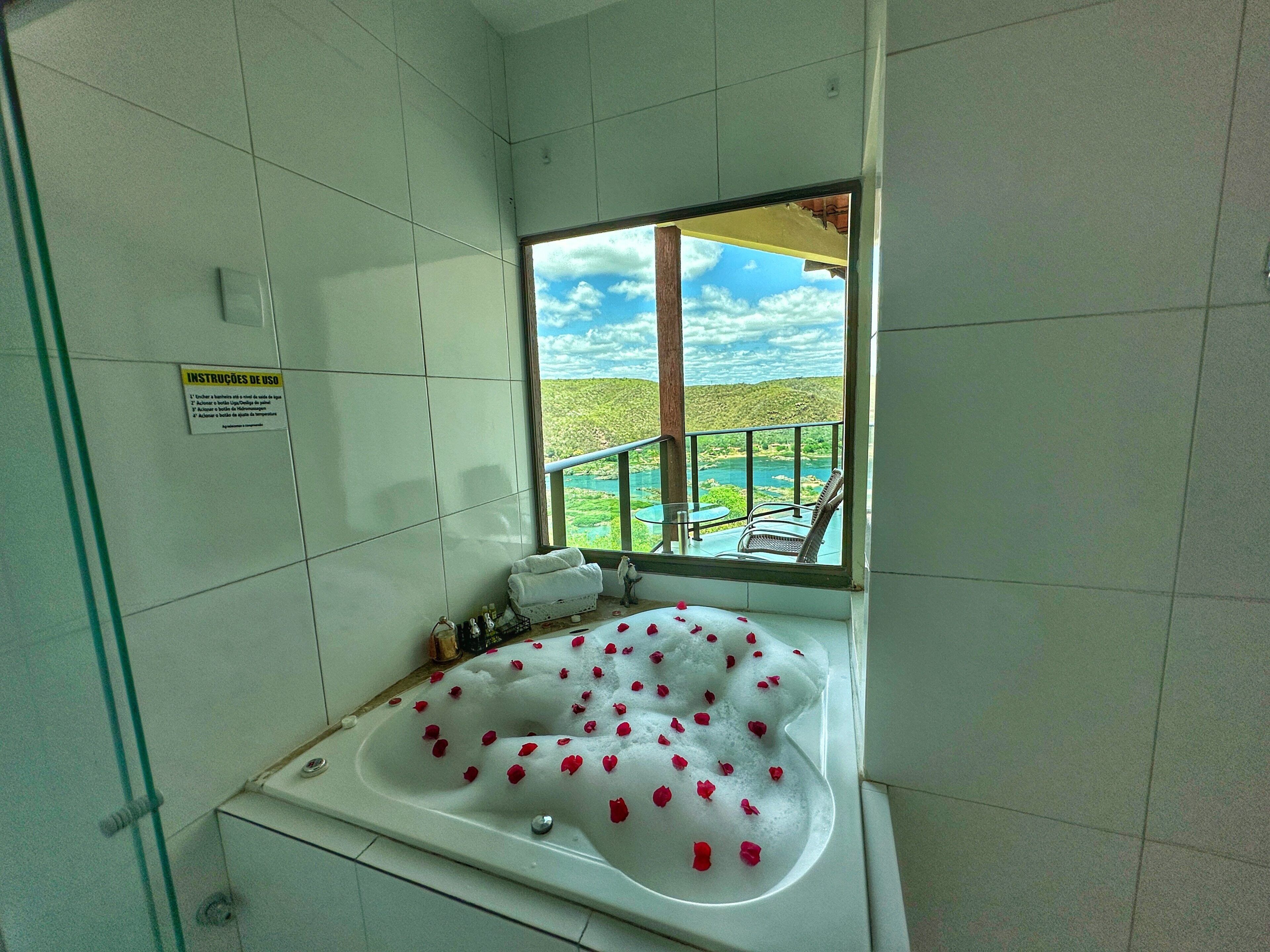 A bathroom with a bathtub filled with bubbles and rose petals, a window with a view of a green landscape, and a sign with usage instructions.