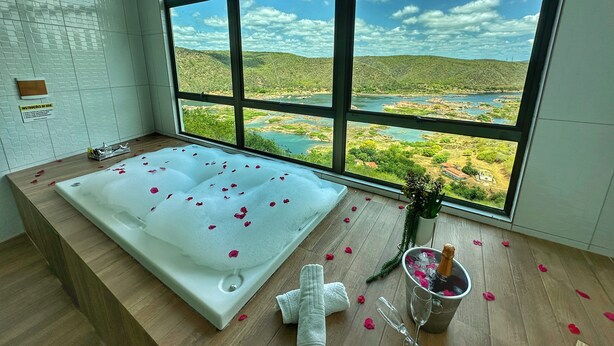 Un baño con una bañera grande, un ventanal con vistas panorámicas y un cubo con una botella y vasos en el suelo.