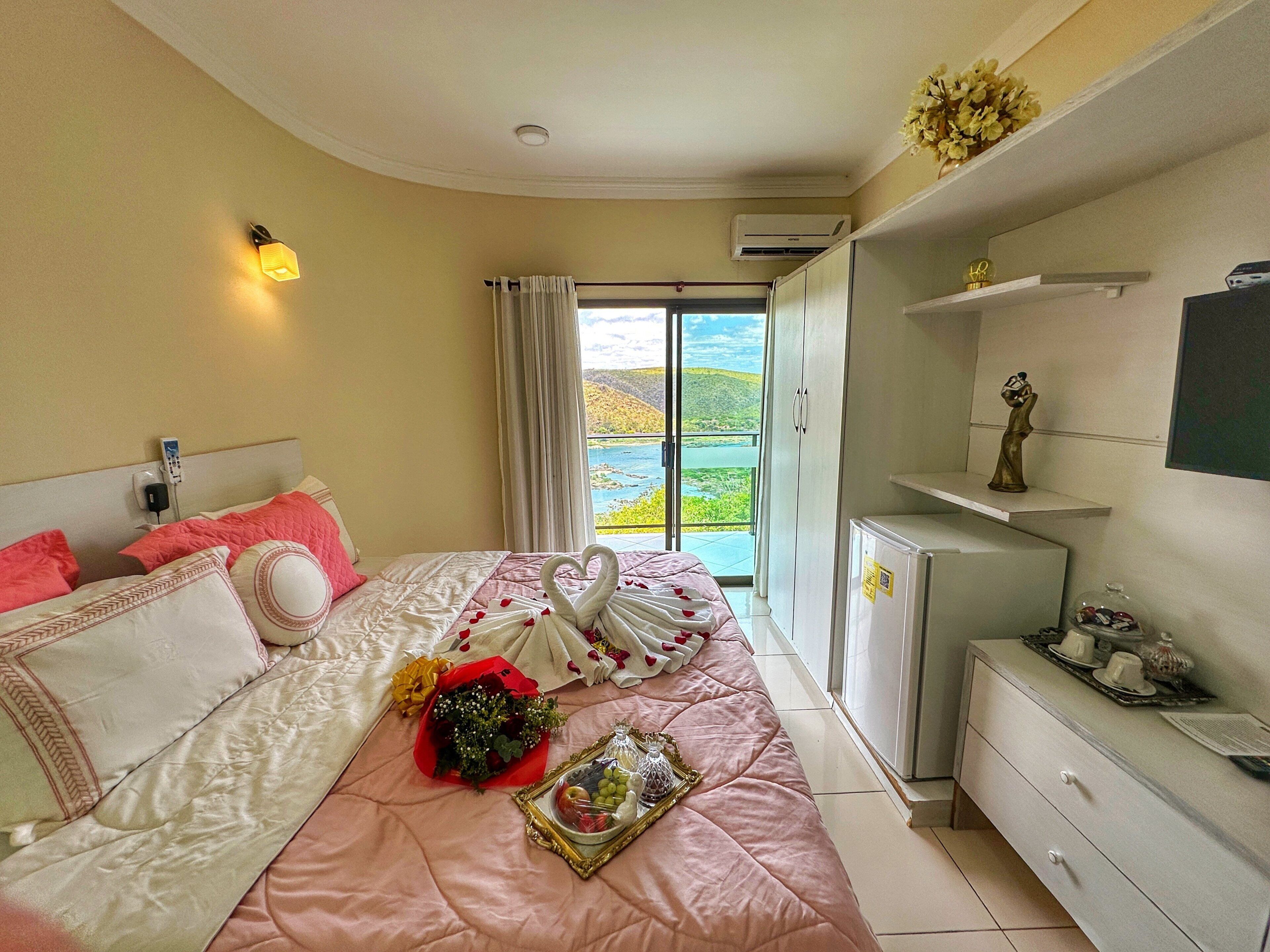 Habitación de hotel con cama, un refrigerador pequeño, un escritorio y vista a un paisaje verde a través de un ventanal grande.