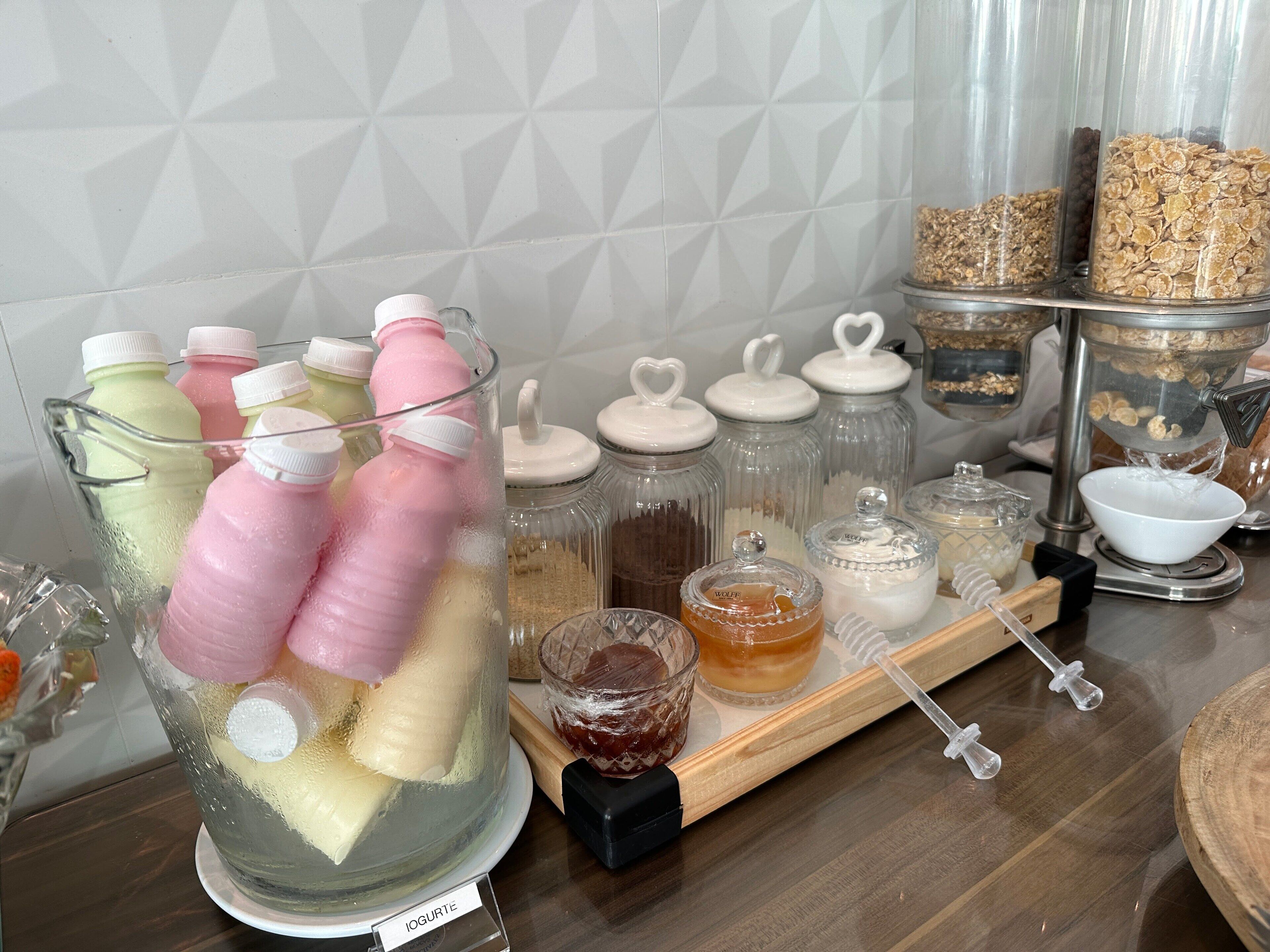 A kitchen counter with various food items and containers, including a large pitcher with bottles, jars with lids, and a wooden board with a honey dipper.