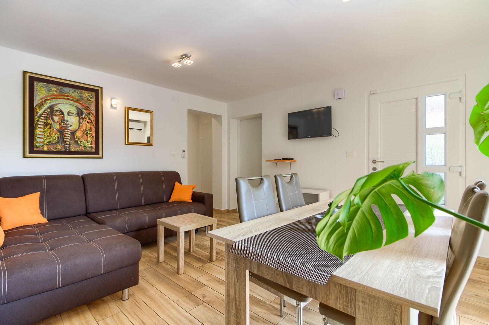 A modern living room with a sofa, coffee table, and dining table. There is a TV mounted on the wall and a large plant in the corner.