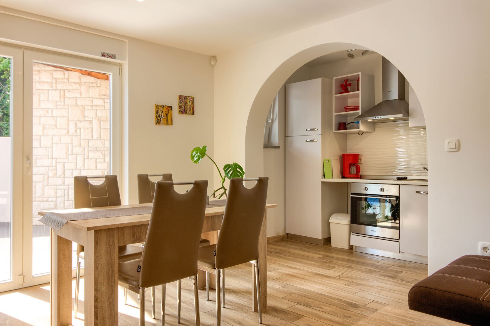 A modern kitchen with an archway, a dining area with a wooden table and chairs, and a view of the outside through a glass door.