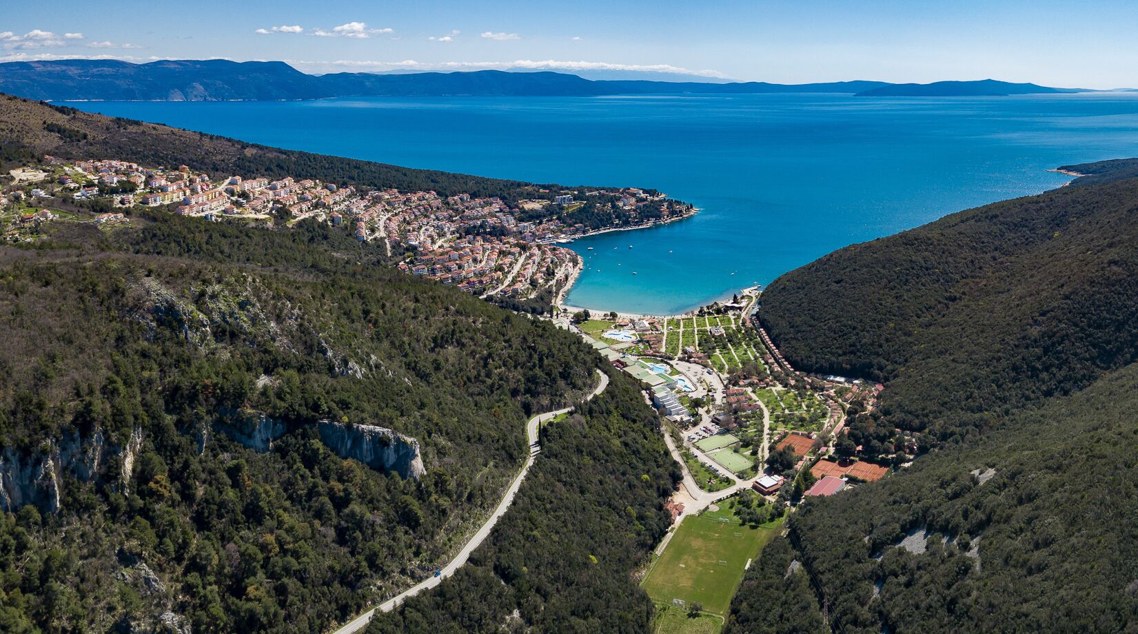 Vue aérienne d’une ville côtière, avec une route sinueuse, une forêt dense et une mer bleue et limpide.
