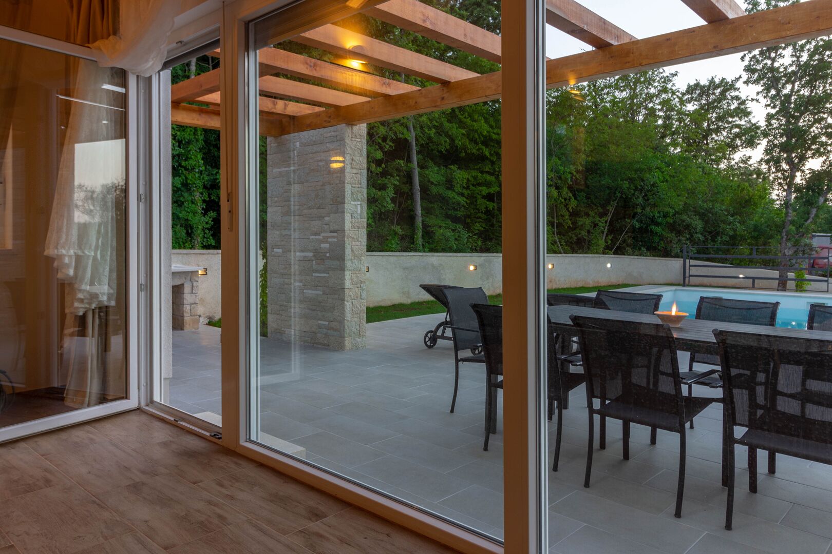 A modern outdoor patio with a glass door leading to a pool area, featuring a dining table and chairs, stone pillars, and wooden beams.