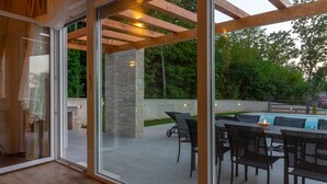 A modern outdoor patio with a glass door leading to a pool area, featuring a dining table and chairs, stone pillars, and wooden beams.