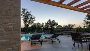 An outdoor pool area with lounge chairs and a table, surrounded by stone walls and trees.