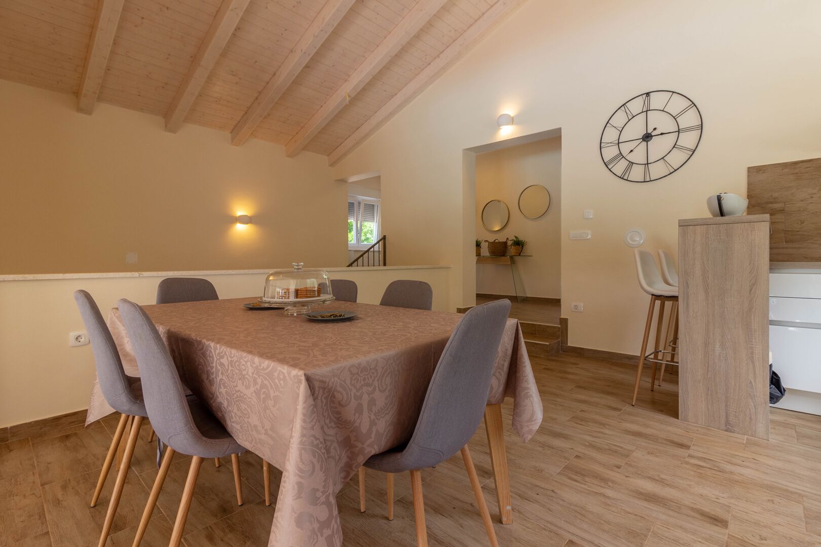 A dining area with a table set for six, a wooden ceiling, and a wall clock.