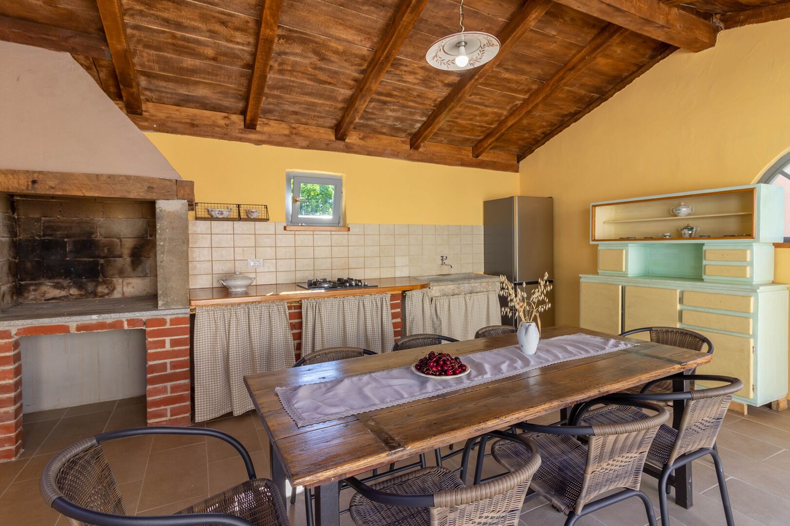 A rustic kitchen with a brick oven, wooden ceiling beams, and a dining area with wicker chairs.