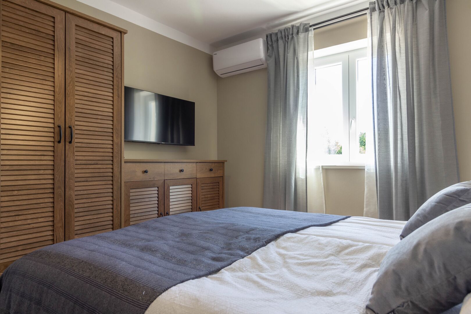 A bedroom with a bed, a TV mounted on the wall, and a window with curtains.