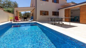 Piscina com espreguiçadeiras, mesa de pingue-pongue e um edifício com várias janelas e porta.