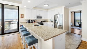 Une cuisine moderne dotée d’un grand îlot central, de meubles blancs, d’appareils électroménagers en acier inoxydable et offrant une vue sur l’extérieur.