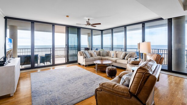 Un salon spacieux avec un grand canapé, une table basse et un téléviseur posé sur un meuble blanc. La pièce est dotée de grandes fenêtres et d’un balcon offrant une vue sur l’océan.