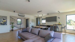 A modern living room with a large L-shaped sofa, a dining area with a wooden table, and a kitchen with a large window. - Villa Esire (Istria - Pula) (Pula)