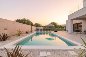 Uma área de piscina ao ar livre moderna, com muro de concreto, espreguiçadeiras e uma pérgola.