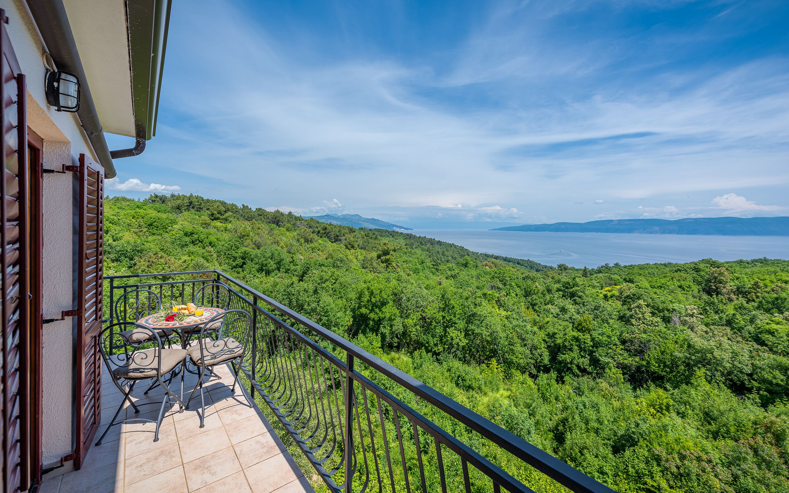 A balcony with a table and chairs, overlooking a lush green landscape and a body of water.