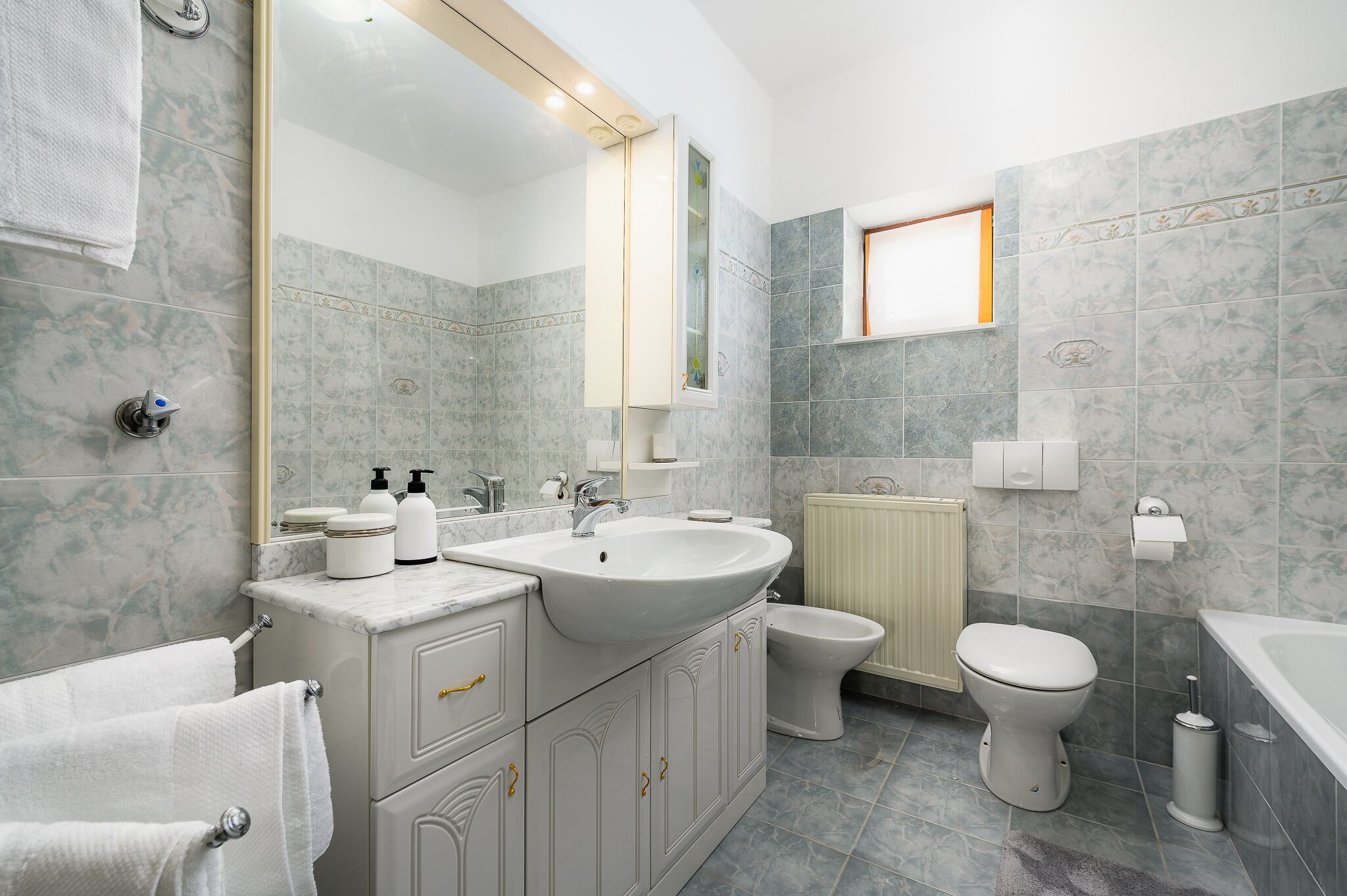 A bathroom with a large sink, toilet, and bathtub. There is a window providing natural light and a towel rack.