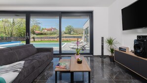 Una sala de estar moderna con un amplio ventanal corredera que da a una zona de piscina, un sofá de cuero oscuro, una mesa de centro de madera con libros y una taza, y un televisor de pantalla plana montado en la pared.
