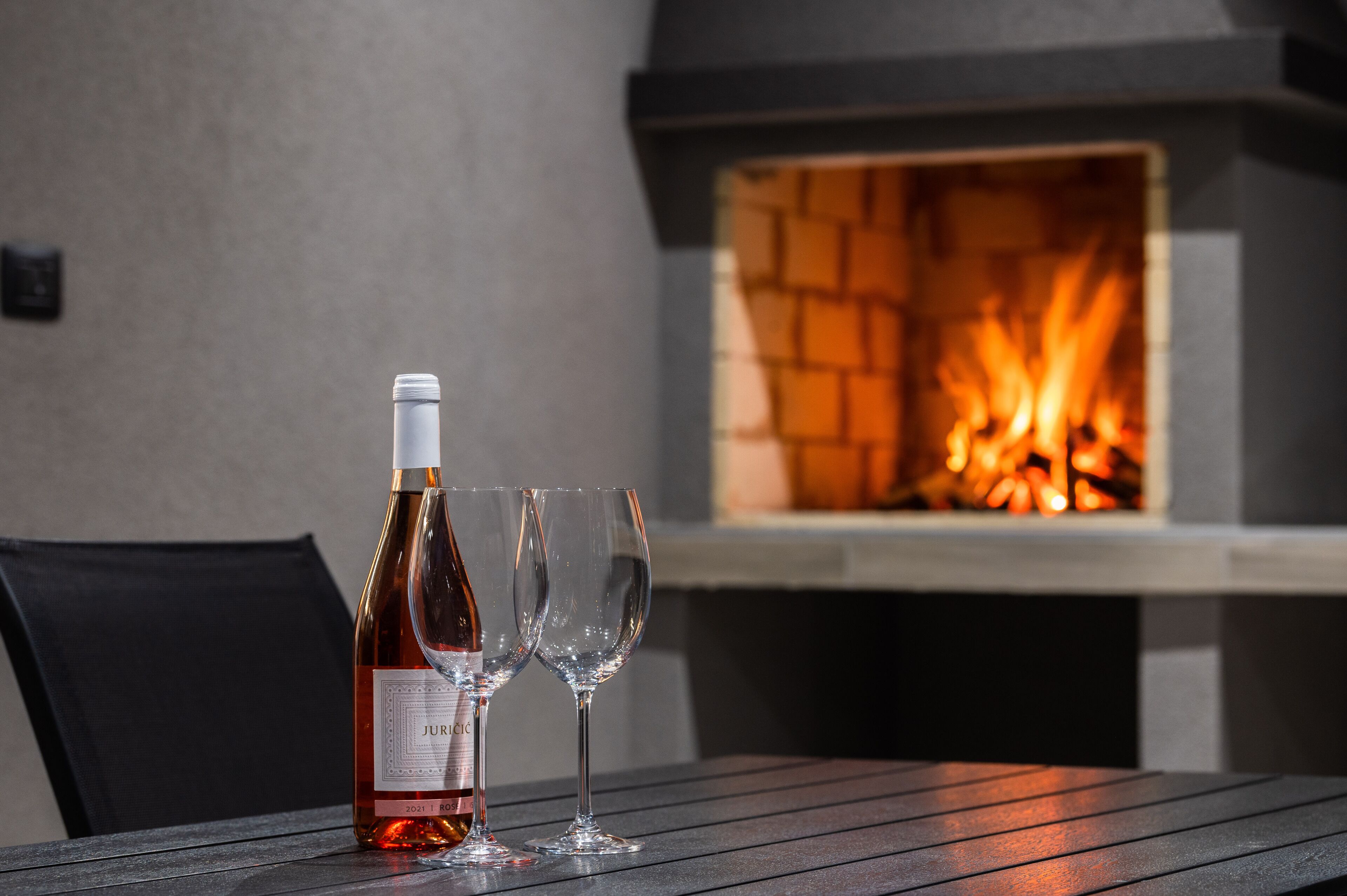 A bottle of wine and two glasses on a table with a fire feature in the background.